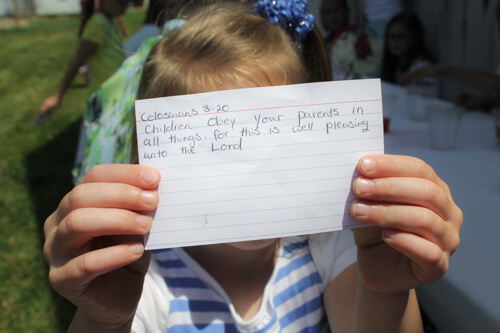A young girl holds up a card with Colossians 3:20 scripture written on it.