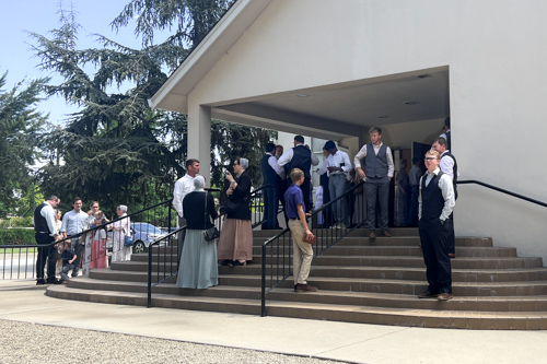 On a sunny day, groups of church members talk with each other outside of a church meeting house.