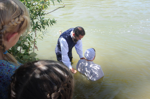 A minister, standing in running water, prepares to baptize a lady kneeling in the water by washing water over her shoulders with his hand.