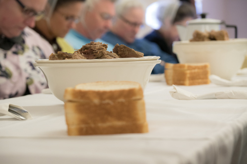 A plate of hot beef sits on a table as church members bow their heads in prayer before the Lord's supper meal.