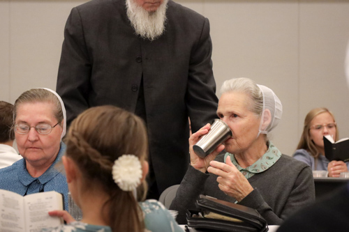 A lady thoughtfully drinks communion wine from a metal cup as a minister watches behind her. Two young girls sing from their hymn books