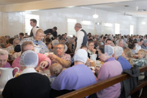 As a large crowd eats a lovefeast lunch in a church, a smiling woman prepares to refill a guest's cup.