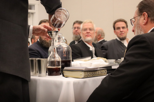 A man pours communion wine from a small glass into a larger jar as several communion participants solemnly watch the process.