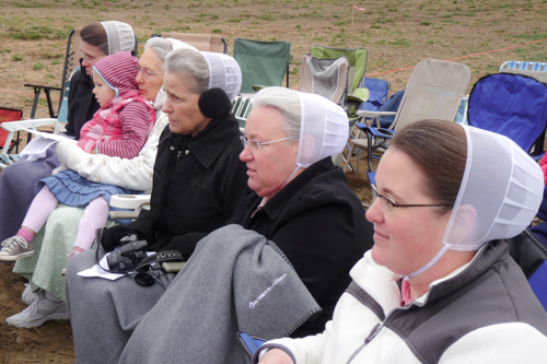 Five women wearing white, see-through head coverings sit outside on camping chairs with blankets and coats.