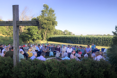 A group of church members in the evening sun sit in a circle around a fire ring as they sing from their songbooks. A wooden cross stands in the foreground in front of the group.