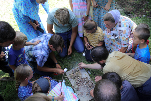 Little boys and girls huddle in a circle on the ground as a few adults try to light a fire for the waiting marshmallows.