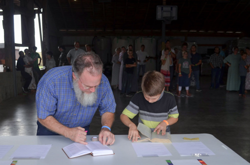 A man and boy lean over a table as they diligently search through some books. A crowd of teenagers and adults in the background look on.