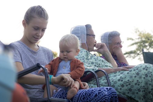 A teenage girl plays with a baby boy in her lap. Two elderly women sitting to the girl's left chat with each toner.