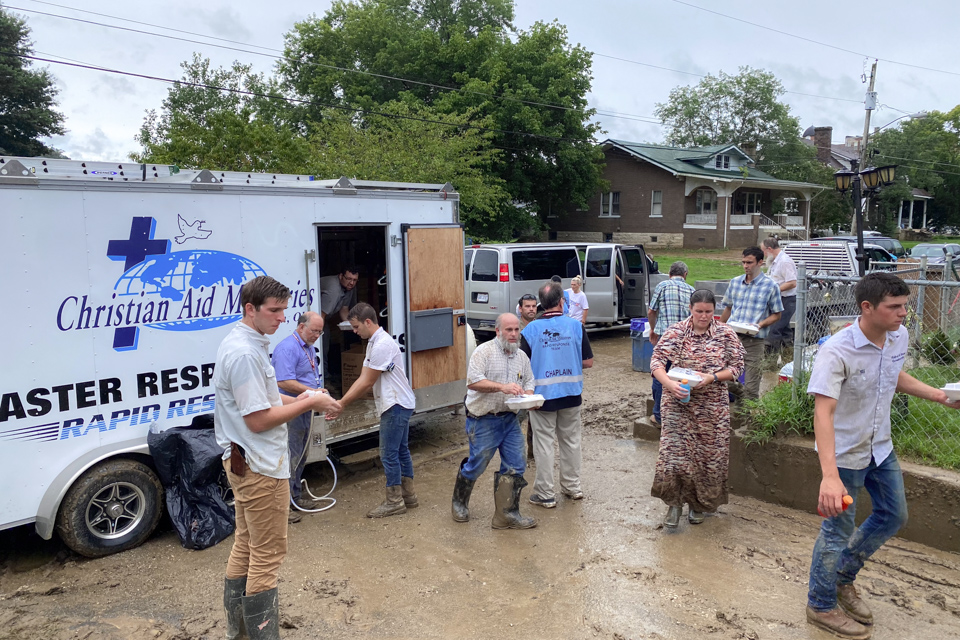 People stand in the muddy driveway beside a Christian Aid Ministries Disaster Response trailer.