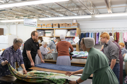 Men and women fold warm comforters together in a room full of clothes and other fabrics.
