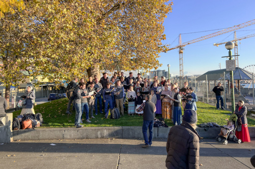 A group of young people sing on a city sidewalk as a passerby looks on.