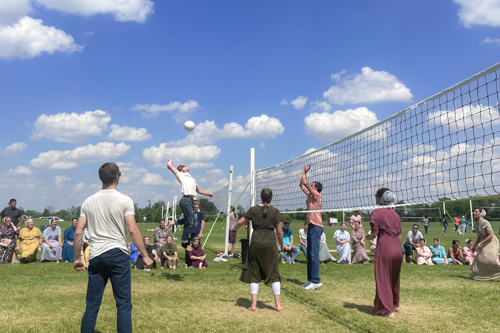 A crowd of young folks line a volleyball court as a young man leaps in the air to spike the volleyball.