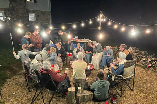 A group of family and friends sit in a tight circle as they chat and roast marshmallows over a fire bowl.