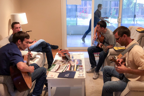 Four young men sitting around a living room play their guitars together.