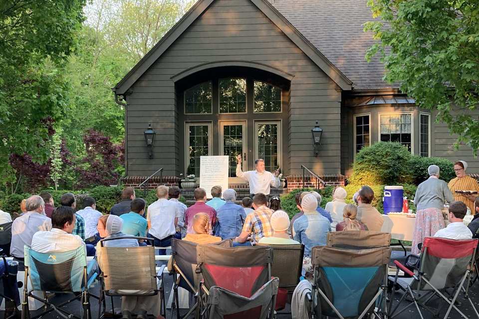 A crowd sits outside on camping chairs as a man standing next to an easel teaches them.