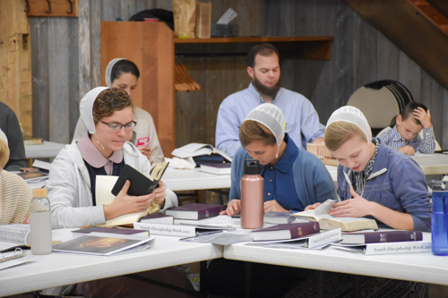 Young ladies sit at a table, searching through their Bibles and taking notes. A family sitting behind them do the same.