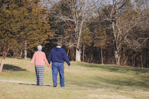 A husband and wife hold hands as they walk through a clearing in the fall woods.