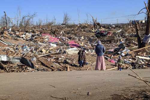A woman and her child talk with a man who kneels in front of a landscape filled with house wreckage and destruction.