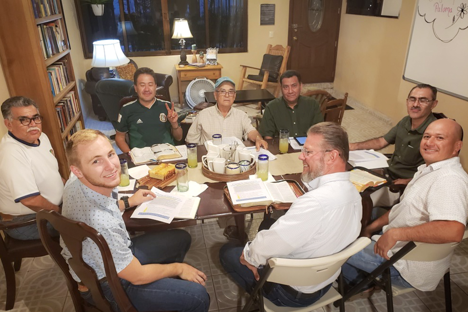 Eight men of various ethnicities sit around a dining room table as they study their Bibles and converse.