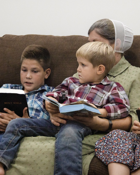 Two boys, one sitting on his mother's lap, sit on a couch as they read their Bibles.
