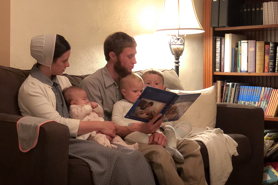 A family sits on a couch in their living room. The father reads a book while his wife and three little children listen and follow along.
