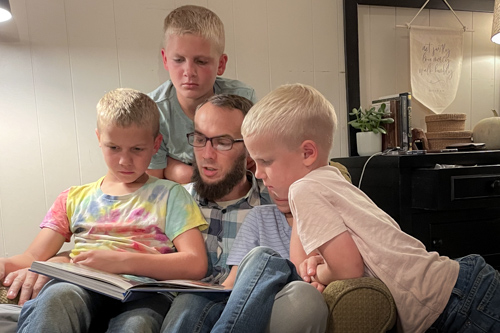 Four young boys gather around their father on a chair as he reads a book to them.