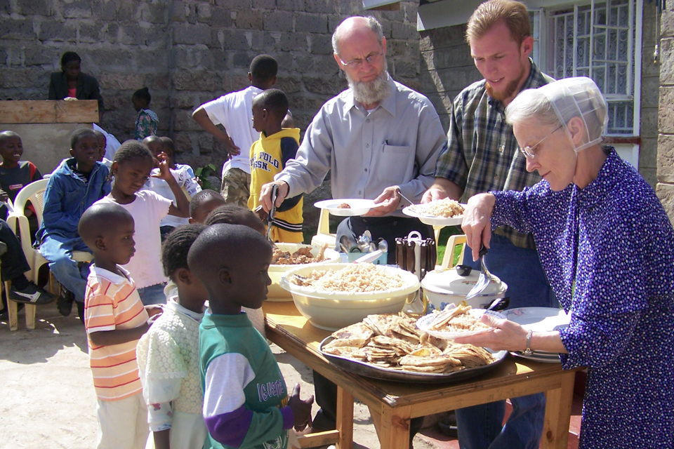 In a sunny courtyard, three missionary works scoop food onto plates for a group of young kids waiting in line.