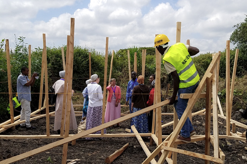 A local man in a hard hat perches on wood framing as he builds a wall. A group of lady missionaries behind him talk with other locals.