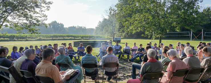 On a bright summer morning, a large group of young people sit around a campfire and read their Bibles.